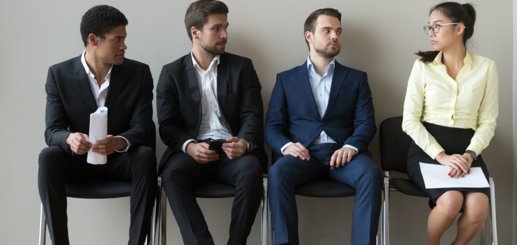 Microagressão: Estão sentados numa cadeira três homens e uma mulher de saia olhando para eles .