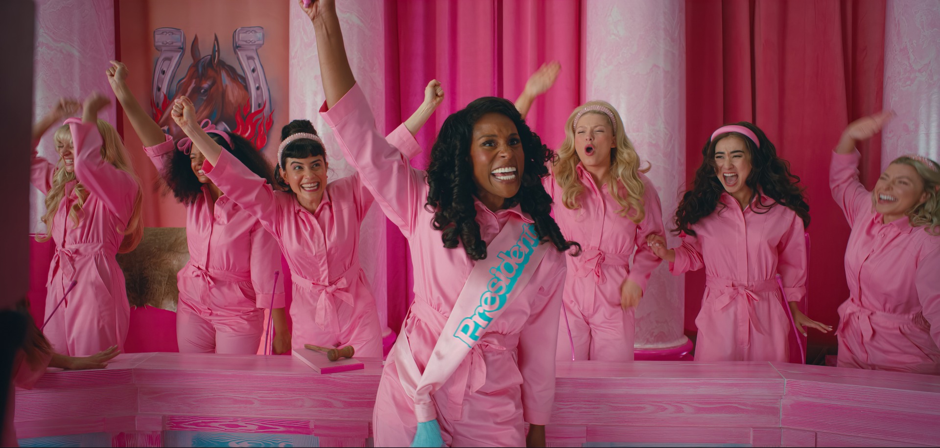 Barbies dançam e comemoram. Todas vestem rosa pink. Uma Barbie negra com faixa de presidente está na frente