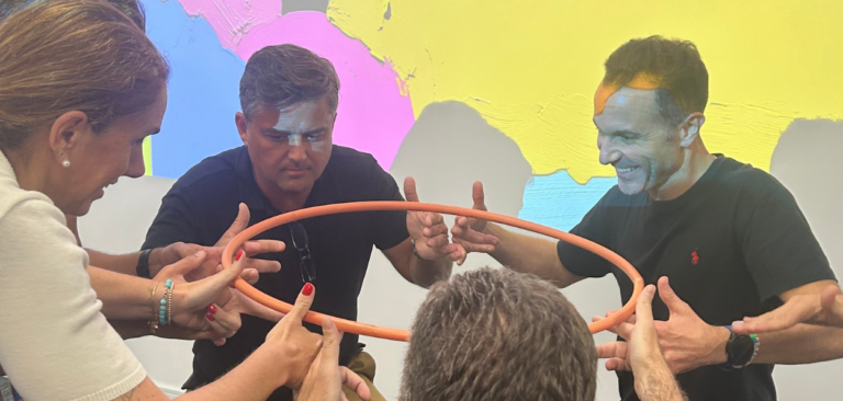 Na imagens algumas pessoas, homens e mulheres, equilibram juntos um bambole usando os dedos indicadores. Eles se divertem e se conscientizam na oficina Carlotas Empresas